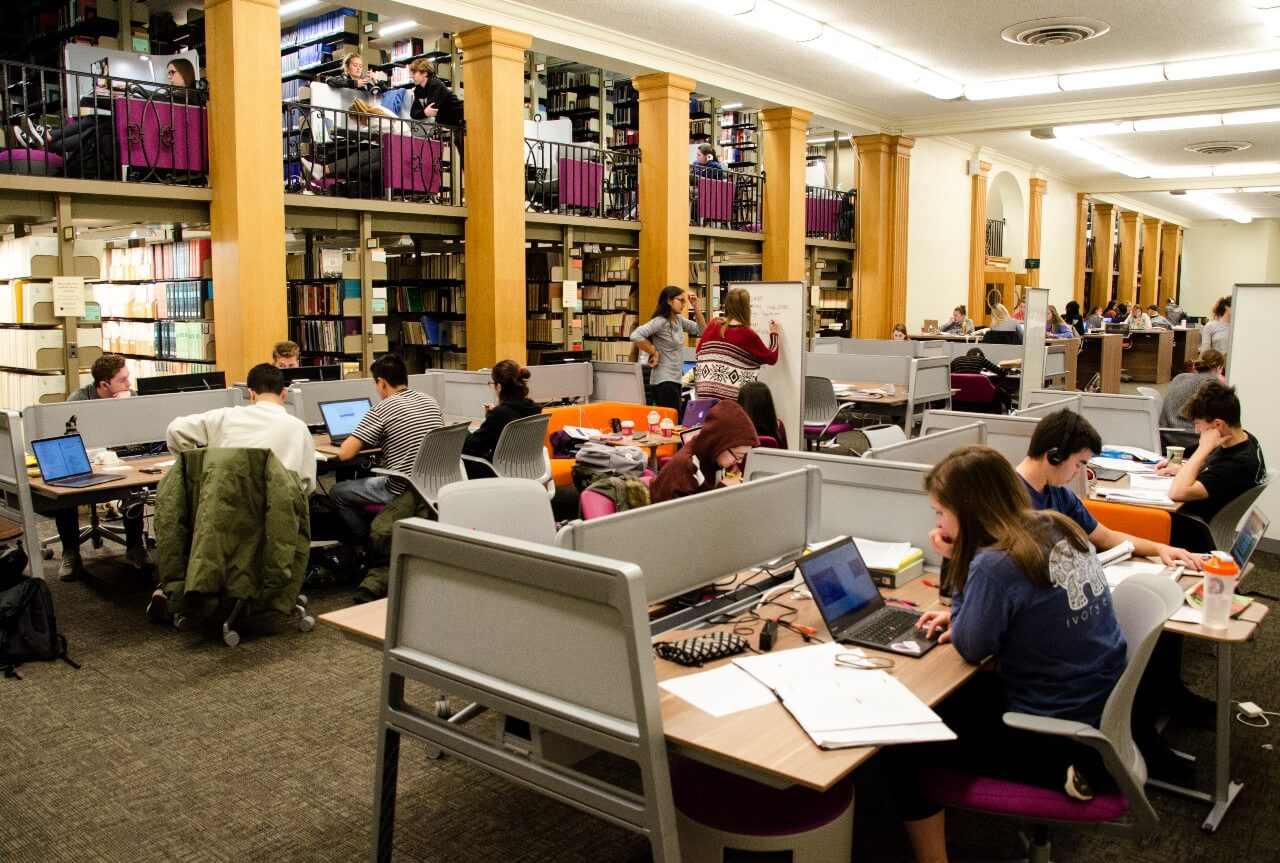 An image of many students in a library study space. Some are sitting at tables, some are standing and writing on whiteboards, some are engaged in conversations.