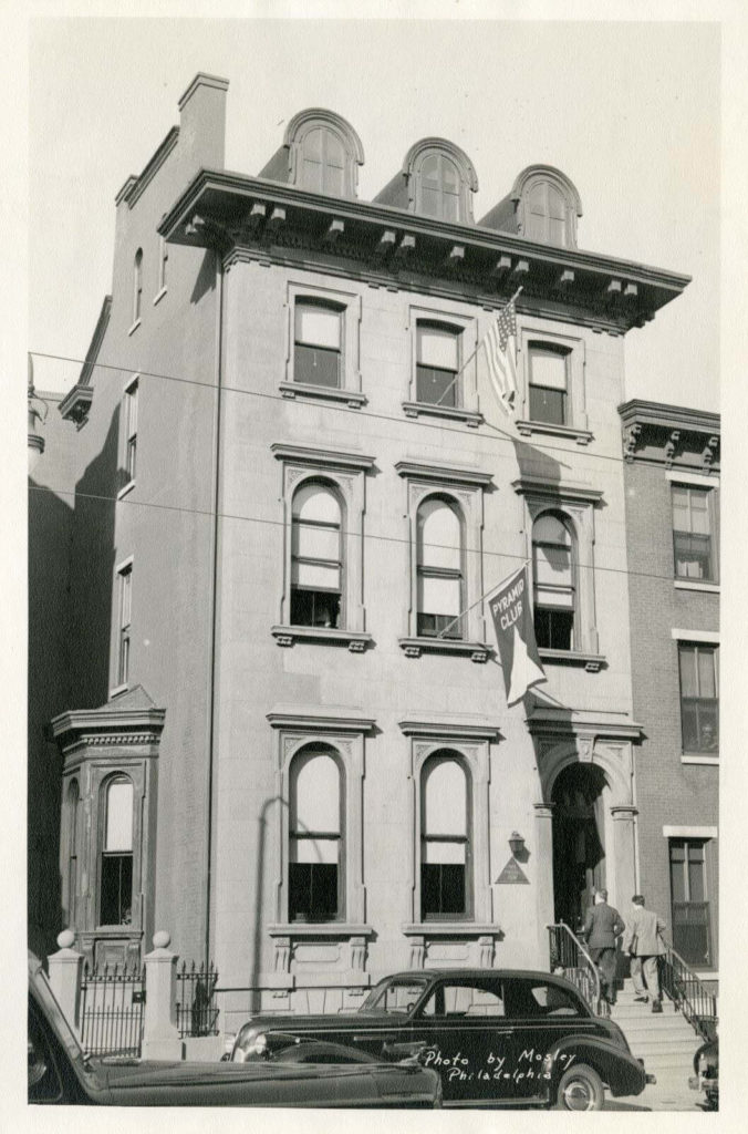 Black and white photo of a 3 story brown-stone row home with an American flag out of the top, middle window and a Pyramid Club flag sticking out of the 2nd story, middle window. There are 2 men walking up stairs into the building and cars parked on the street.