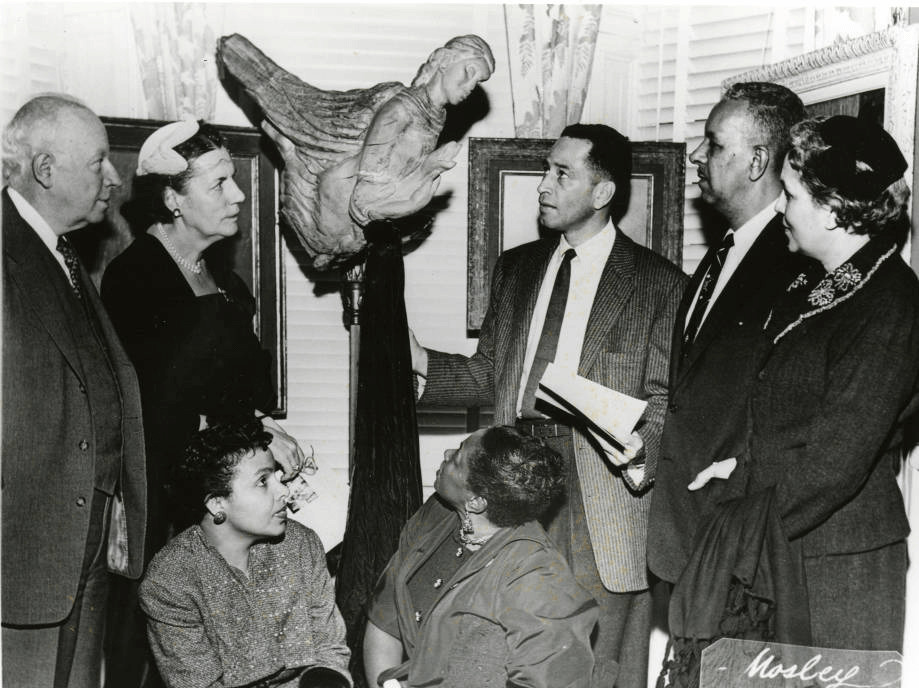 Humbert Howard with Selma Burke, Hale Woodruff, and 4 other people surrounding Burke's Dancing Angels sculpture. With framed paintings behind them, these people surround a statue of the torso of an angel, with praying hands, mounted on a tall pillar that's taller than the standing people.